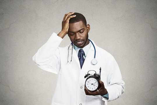 Closeup portrait overwhelmed with busy schedule health care professional doctor, nurse, dentist with stethoscope holding alarm clock running out of time having headache isolated black grey background
